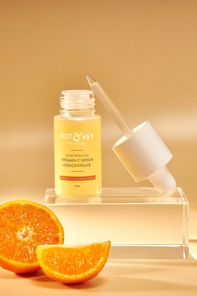 A high-clarity product photography shot of a Vitamin C serum bottle placed on a clear acrylic block, surrounded by fresh orange slices.